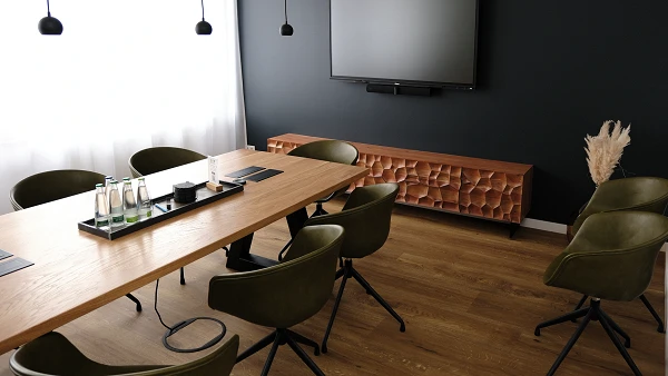 Ein modernes Besprechungszimmer mit einem langen Holztisch, umgeben von grünen Stühlen. An der Wand hängt ein großer Fernseher, und auf dem Tisch stehen Wasserflaschen sowie ein dekoratives Element. Der Raum hat eine dunkle Wandfarbe und viel natürliches Licht.