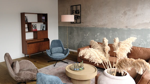 Ein modern gestalteter Aufenthaltsraum mit komfortablen Sitzgelegenheiten in sanften Farben, einer Holzkommode mit Bücherregal, einer Stehlampe und einem runden Couchtisch, umgeben von Wänden, die einen Kombination aus glatten und strukturierten Oberflächen zeigen. Grønne Pflanzen und dekorative Elemente ergänzen die Einrichtung.