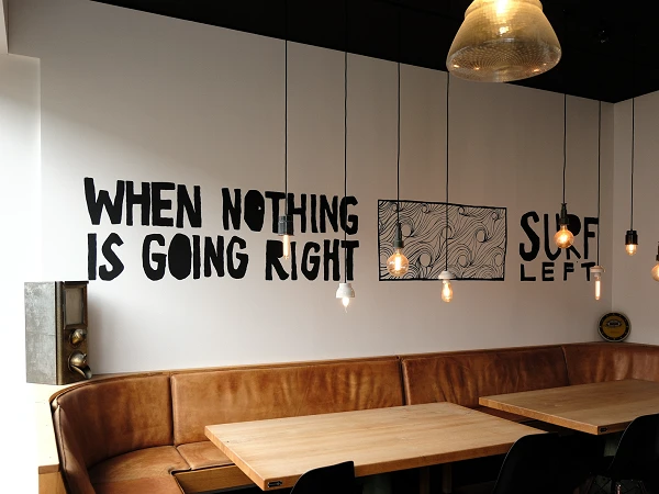 Der Innenraum eines Cafés oder Restaurants mit einer gemütlichen Sitzbank an der Wand und einem großen Holztisch in der Mitte. An der Wand steht der Schriftzug „WHEN NOTHING IS GOING RIGHT SURE LEFT“ in auffälliger Schrift. Über den Tischen hängen mehrere dekorative Lampen.