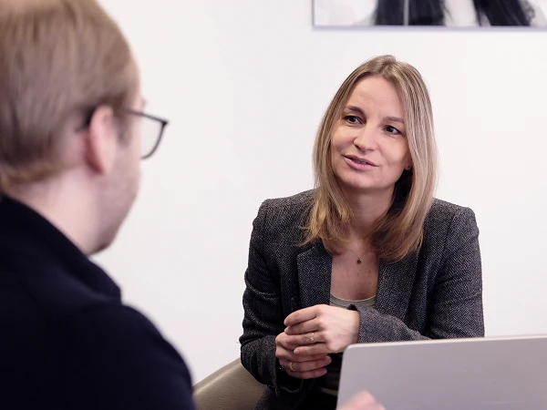 Eine blonde Frau in einem grauen Blazer spricht mit einem Mann, der von der Kamera abgewandt ist, während sie an einem Tisch sitzt, auf dem ein Laptop steht.