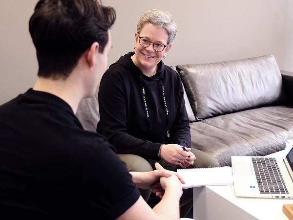Eine Frau mit kurzen grauen Haaren und Brille sitzt auf einem Sofa und lächelt während eines Gesprächs mit einem Mann, der ihr gegenüber sitzt und einen Notizblock in den Händen hält. Auf einem Tisch neben ihnen steht ein Laptop.
