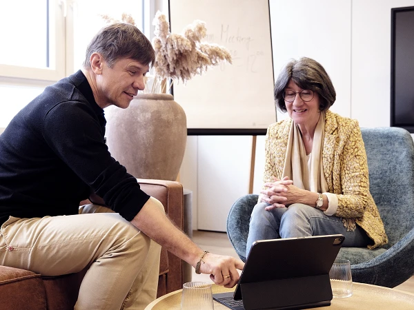Zwei Personen sitzen in einem gemütlichen Raum, während sie an einem Tablet arbeiten; der Mann zeigt auf den Bildschirm, während die Frau aufmerksam zuschaut. Im Hintergrund ist ein Flipchart mit der Aufschrift "Meeting" und eine große Vase mit Trockenblumen zu sehen.