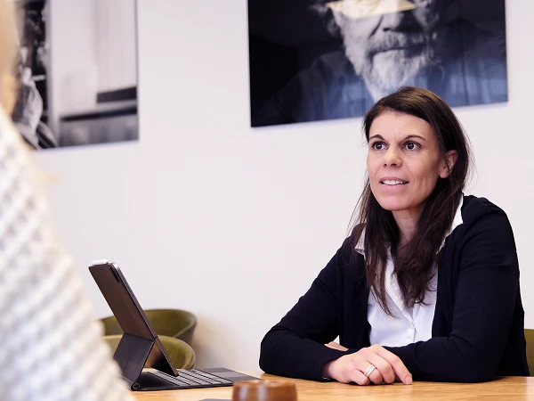 Eine Frau mit langen, dunklen Haaren sitzt an einem Tisch und diskutiert, während sie eine andere Person ansieht. Im Hintergrund sind schwarz-weiße Porträtfotos an der Wand zu sehen, und auf dem Tisch steht ein Tablet.