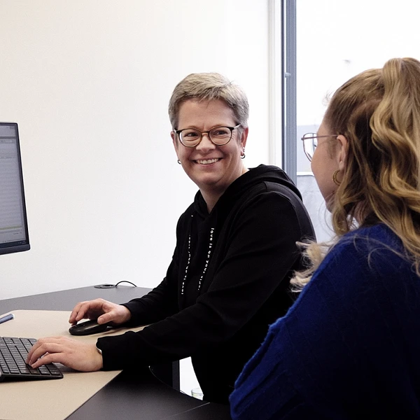 Zwei Frauen sitzen an einem Stehpult und arbeiten gemeinsam am Computer, während sie sich unterhalten. Auf dem Bildschirm sind Tabellenkalkulationen zu sehen. Im Hintergrund sind große Fenster mit Blick auf eine grüne Hecke.