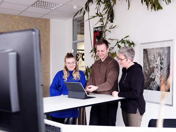 Drei Personen stehen an einem Tisch und betrachten gemeinsam ein Tablet. Im Hintergrund sind Pflanzen und ein Bild an der Wand sichtbar.