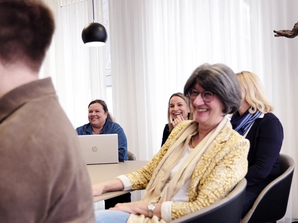 In einem modernen Büroraum sitzen fünf Personen an einem Tisch, die entspannt lachen und miteinander interagieren. An der Wand hängt ein Porträt eines älteren Mannes. Jeder Teilnehmer hat einen Laptop vor sich. Die Atmosphäre wirkt freundlich und informell.