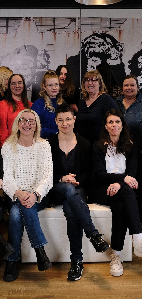 Eine Gruppe von 21 Personen, die in einem modernen Büro steht und sitzt, posiert lächelnd vor einer muralen Wand mit einem künstlerischen Aufdruck. Die Personen sind unterschiedlich gekleidet und scheinen eine diverse Gruppe zu sein.