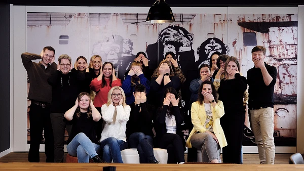 Eine Gruppe von 22 Menschen steht und sitzt in einem Raum, während sie ihre Gesichter mit den Händen verdecken. Im Hintergrund ist eine große Wand mit einem grafischen Wanddesign zu sehen.