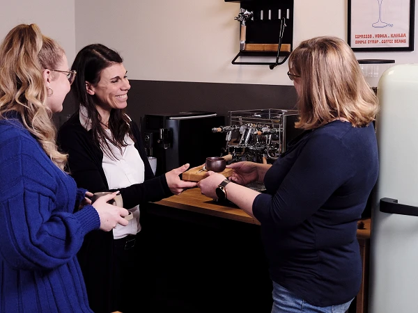 Drei Frauen stehen in einer modernen Küche. Eine Frau überreicht einer anderen einen Becher, während eine dritte Frau lächelnd zuschaut. Im Hintergrund sind eine Kaffeemaschine und ein Kühlschrank zu sehen.