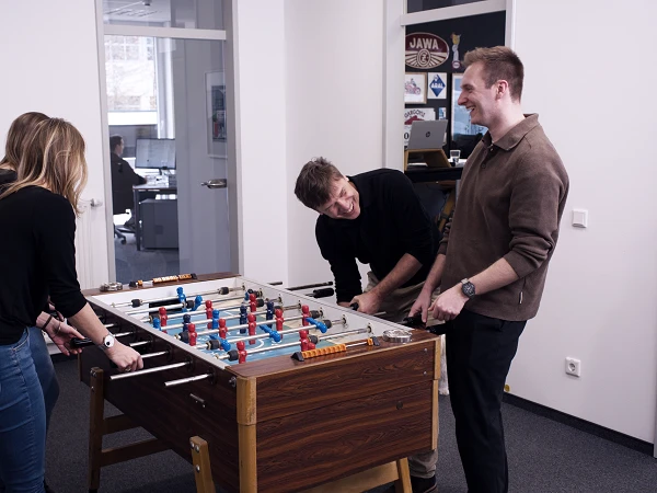 Vier Personen spielen gemeinsam an einem Tischfußballtisch in einem modernen Büro. Zwei Frauen und zwei Männer stehen um den Tisch herum und lächeln, während sie aktiv am Spiel teilnehmen. Im Hintergrund sind Arbeitsplätze und ein Computer sichtbar.