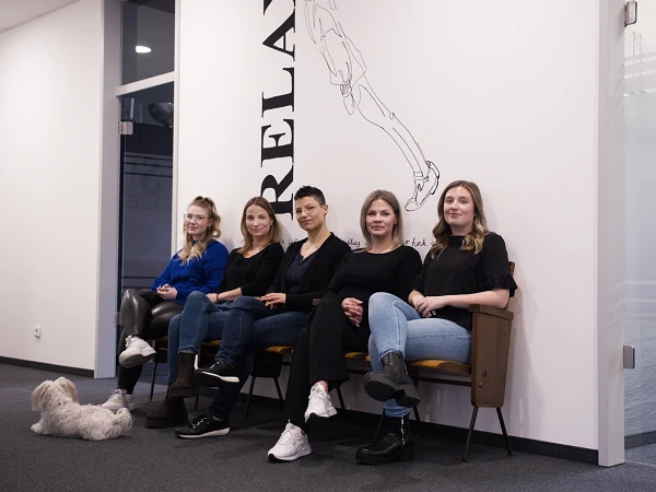 Fünf Frauen sitzen an einer Wand mit Wandgrafik, während ein kleiner Hund auf dem Boden liegt; alle tragen schwarze Oberteile und lächeln in die Kamera.