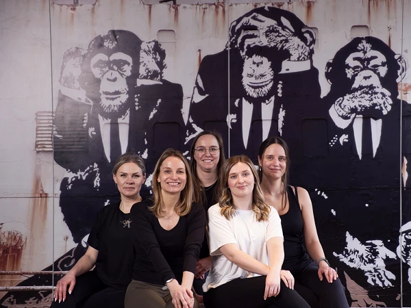 Sechs Frauen posieren in lässiger Kleidung vor einer Wand mit einem großen, grafischen mural von Affen in Anzügen.