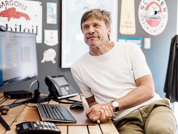 Ein Mann mit kurzem, braunem Haar sitzt entspannt an einem Holztisch, lächelt und schaut zur Seite. Vor ihm stehen ein Computerbildschirm und eine Telefontastatur, während im Hintergrund verschiedene bunte Wanddekorationen zu sehen sind.