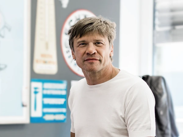 Ein Mann mit kurzen, grauen Haaren trägt ein weißes T-Shirt und lächelt, während er in einem hellen Raum steht. Im Hintergrund sind verschiedene bunte Schilder und ein Thermometer zu sehen.