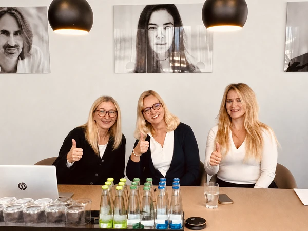 Drei Frauen sitzen an einem Tisch in einem Konferenzraum und zeigen Daumen hoch. Auf dem Tisch stehen Wasserflaschen und Gläser, sowie ein Laptop. Im Hintergrund hängen schwarz-weiße Porträts an der Wand.