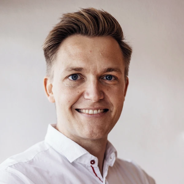 Ein lächelnder Mann mit blonden Haaren und einem weißen Hemd steht mit verschränkten Armen vor einer hellen Wand.