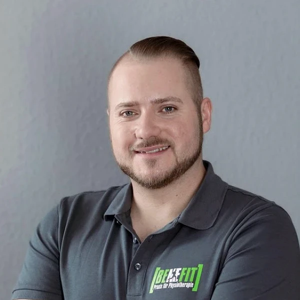 Ein lächelnder Mann mit kurzen, braunen Haaren und einem gepflegten Bart steht mit verschränkten Armen vor einer grauen Wand. Er trägt ein dunkelgraues Polo-Shirt mit dem Logo "Be-Fit" darauf.