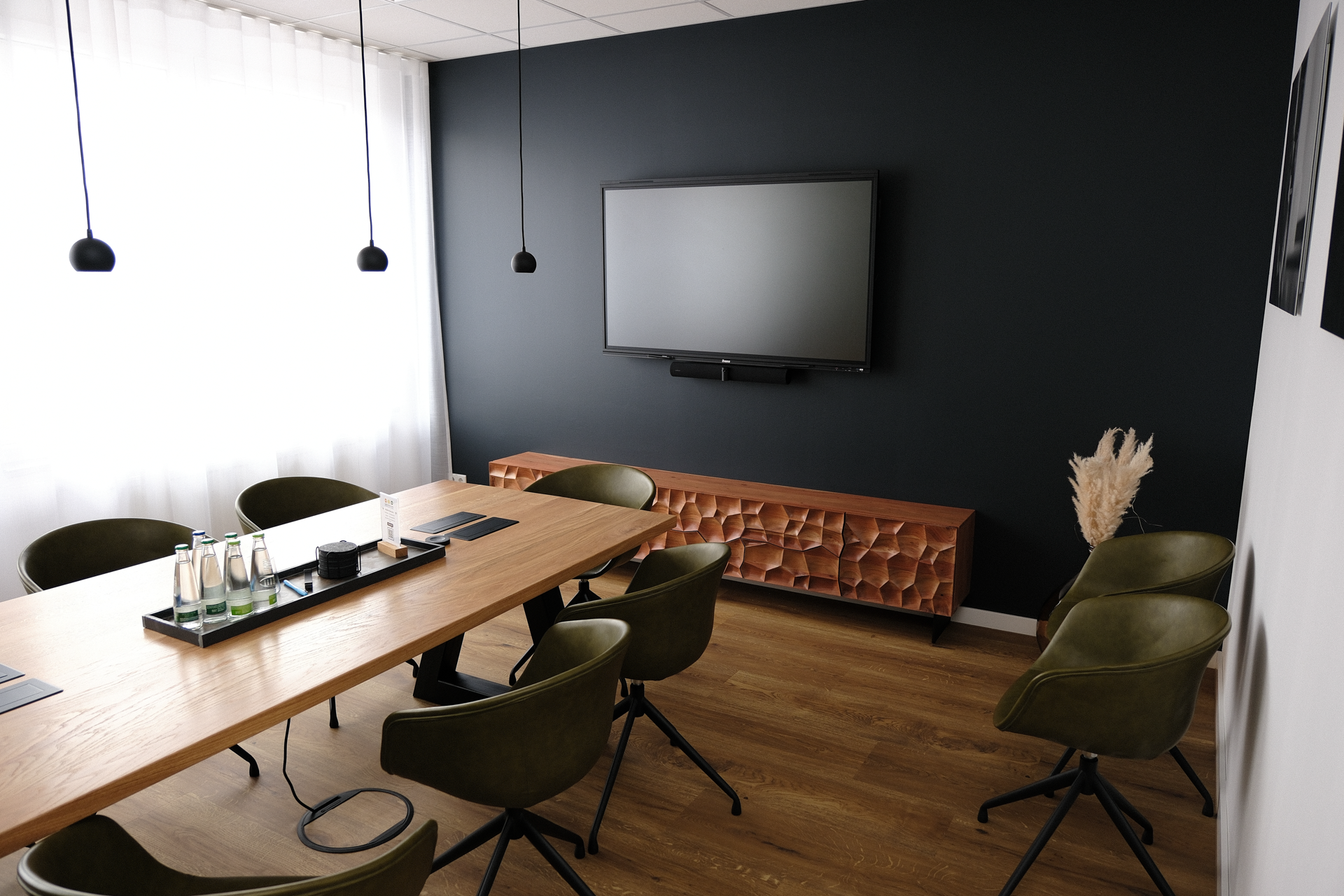 Ein modernes Besprechungszimmer mit einem langen Holztisch, umgeben von grünen Stühlen. An der Wand hängt ein großer Fernseher, und auf dem Tisch stehen Wasserflaschen sowie ein dekoratives Element. Der Raum hat eine dunkle Wandfarbe und viel natürliches Licht.