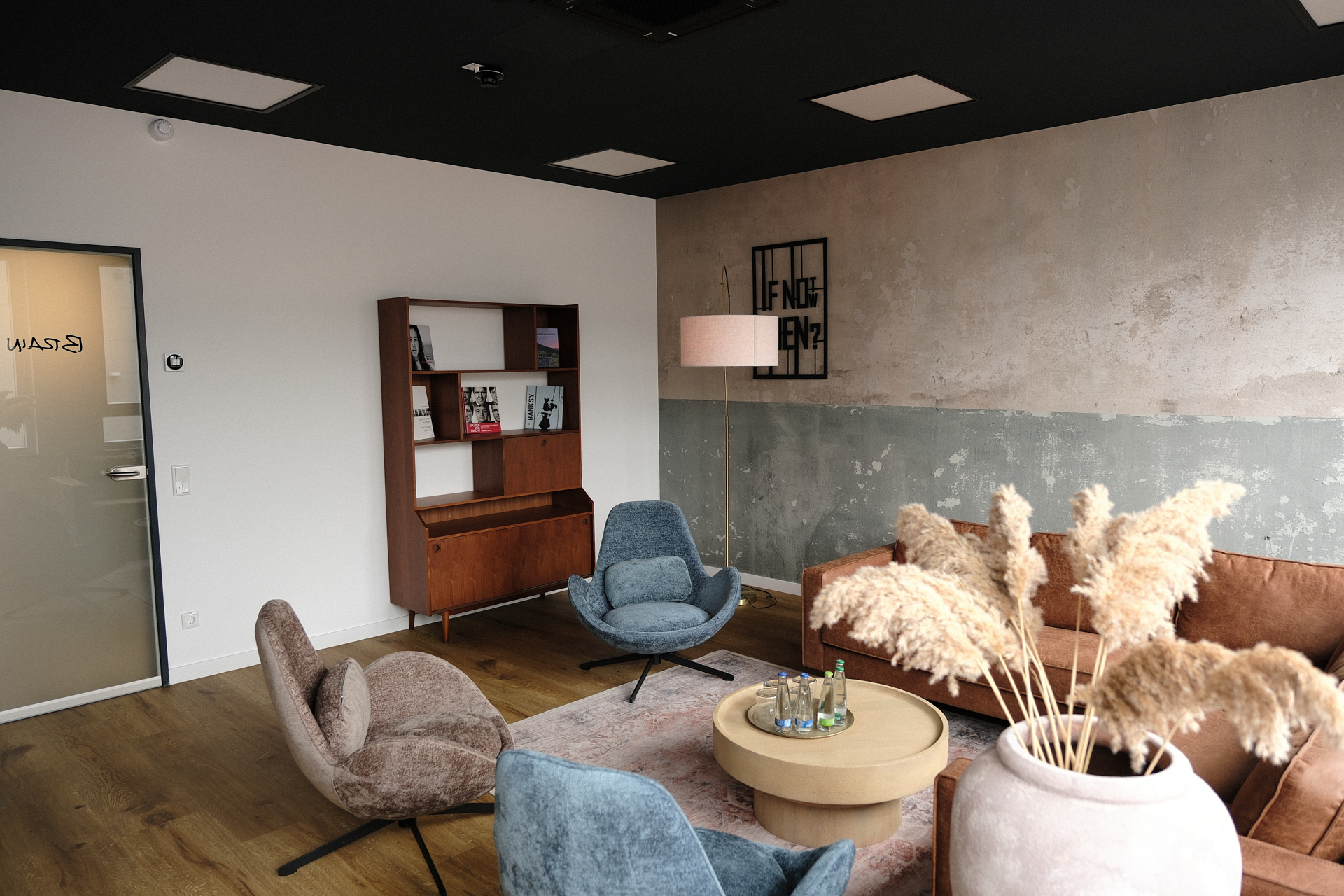 Ein modern gestalteter Aufenthaltsraum mit komfortablen Sitzgelegenheiten in sanften Farben, einer Holzkommode mit Bücherregal, einer Stehlampe und einem runden Couchtisch, umgeben von Wänden, die einen Kombination aus glatten und strukturierten Oberflächen zeigen. Grønne Pflanzen und dekorative Elemente ergänzen die Einrichtung.
