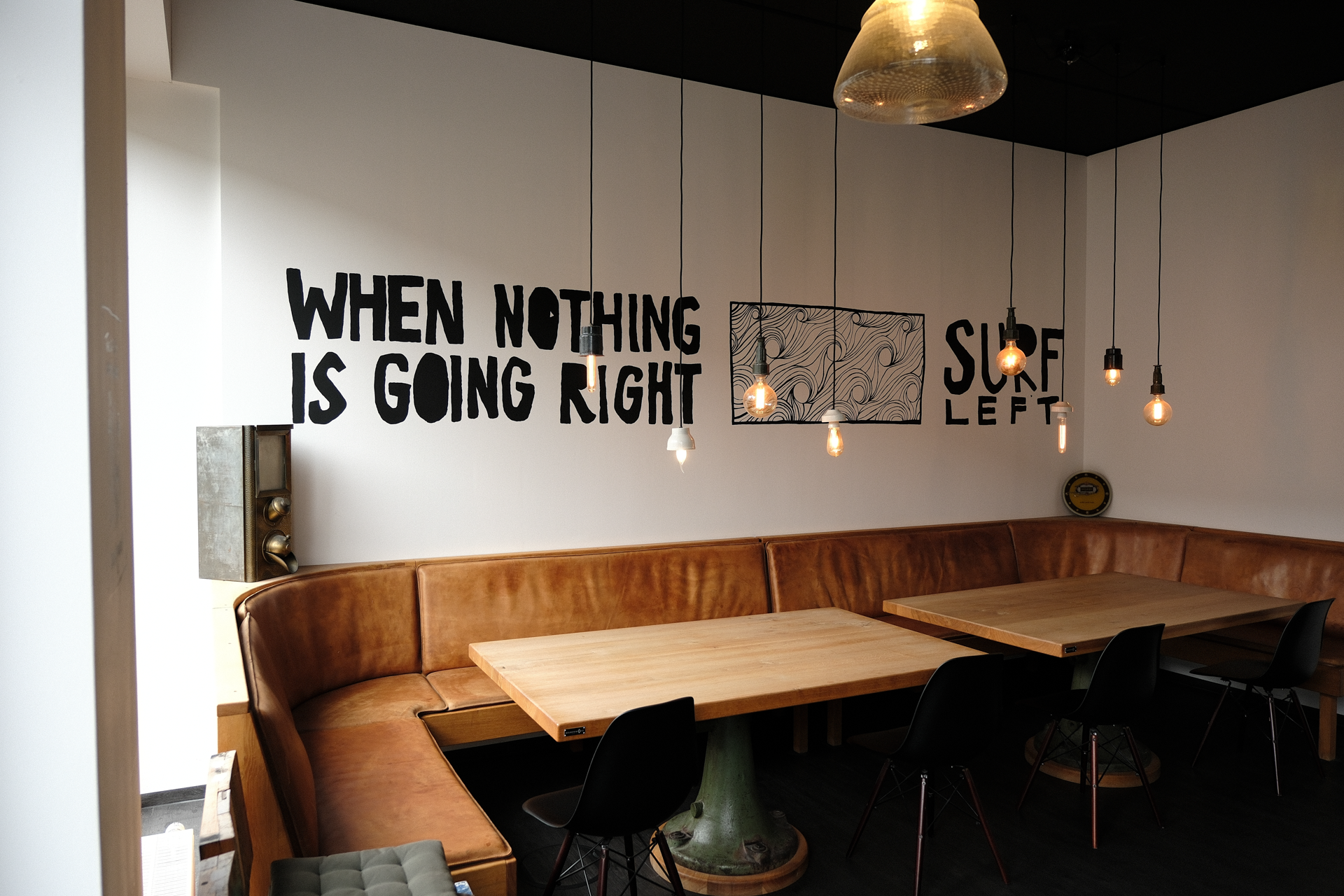 Der Innenraum eines Cafés oder Restaurants mit einer gemütlichen Sitzbank an der Wand und einem großen Holztisch in der Mitte. An der Wand steht der Schriftzug „WHEN NOTHING IS GOING RIGHT SURE LEFT“ in auffälliger Schrift. Über den Tischen hängen mehrere dekorative Lampen.