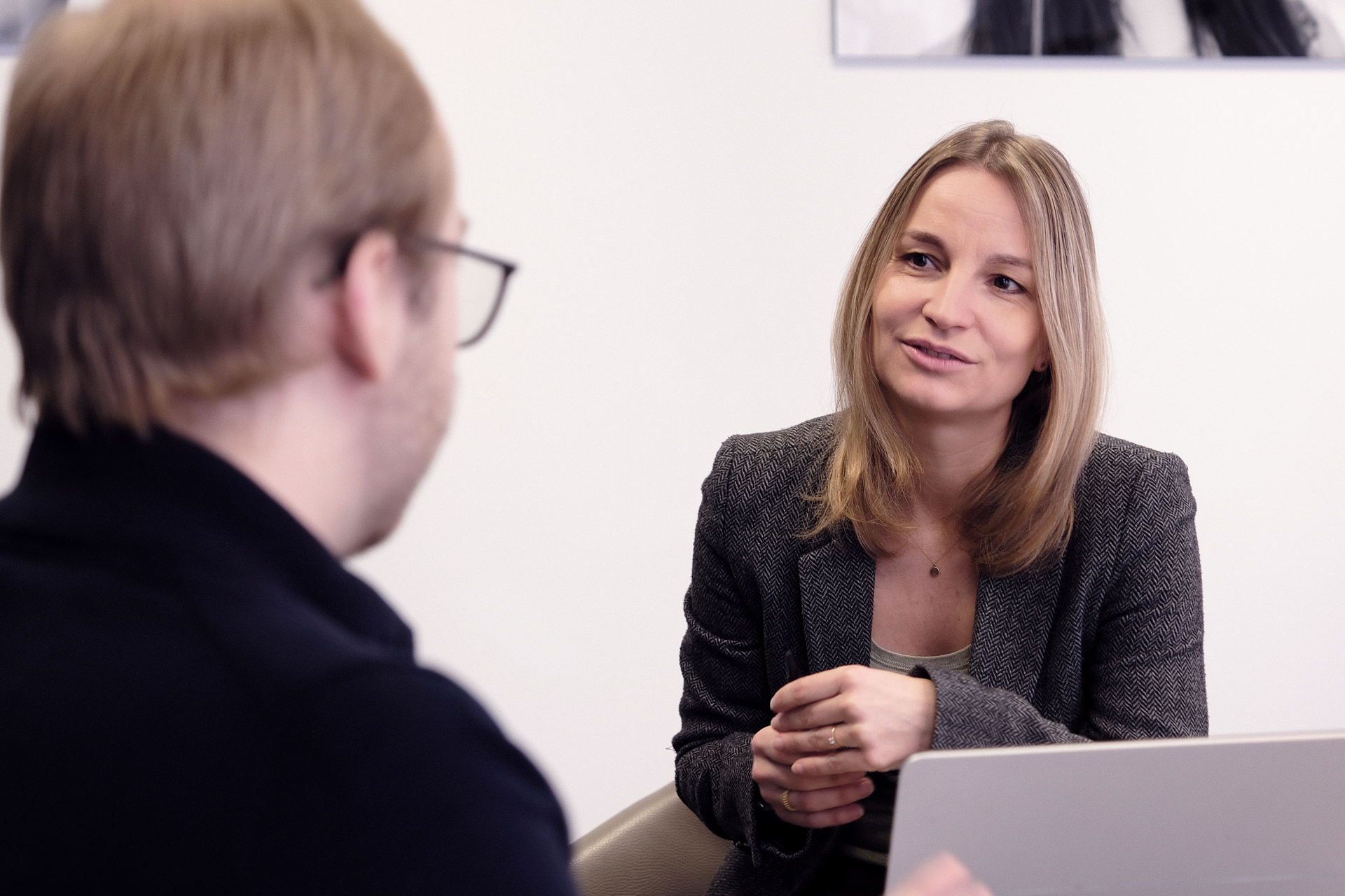 Eine blonde Frau in einem grauen Blazer spricht mit einem Mann, der von der Kamera abgewandt ist, während sie an einem Tisch sitzt, auf dem ein Laptop steht.