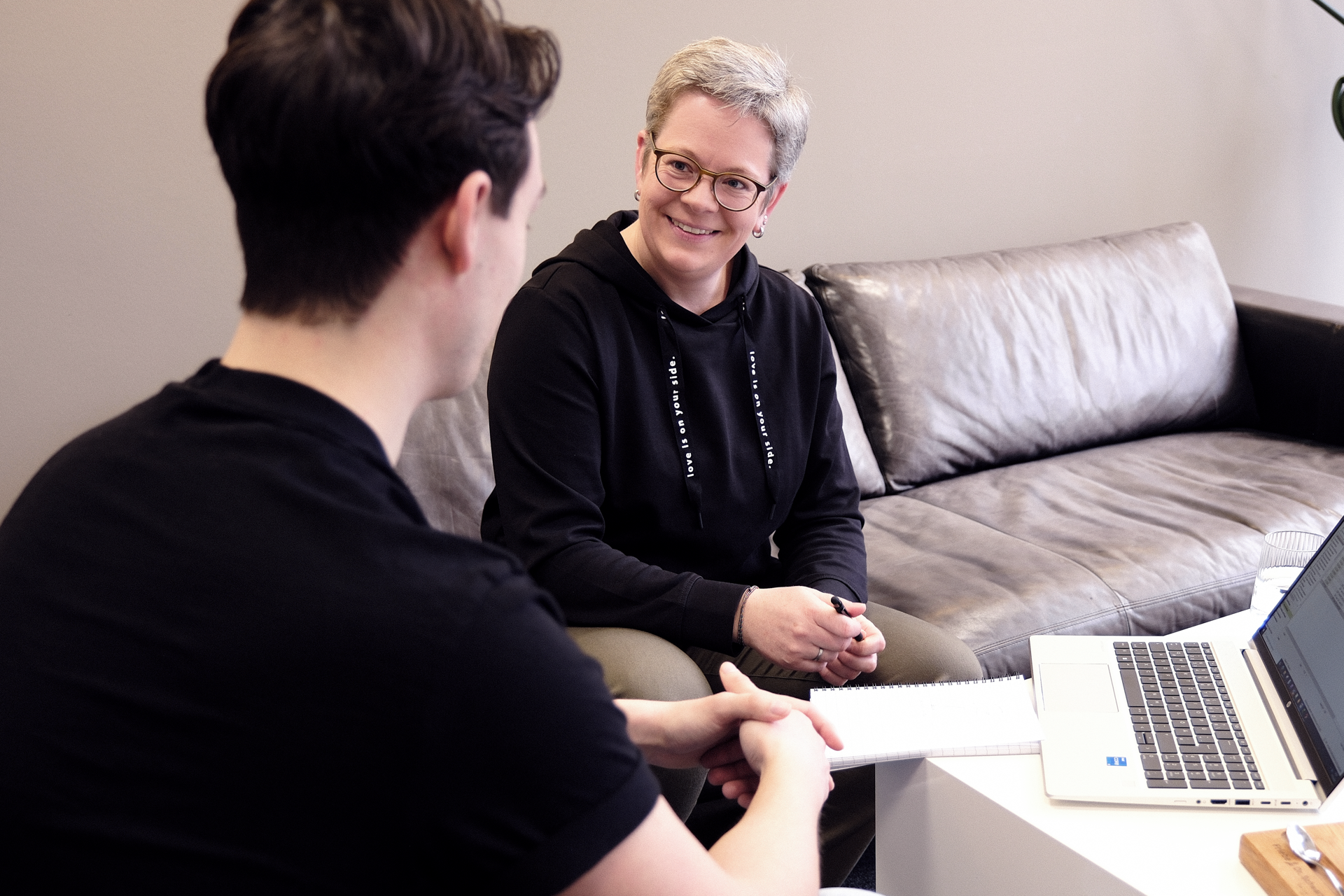 Eine Frau mit kurzen grauen Haaren und Brille sitzt auf einem Sofa und lächelt während eines Gesprächs mit einem Mann, der ihr gegenüber sitzt und einen Notizblock in den Händen hält. Auf einem Tisch neben ihnen steht ein Laptop.