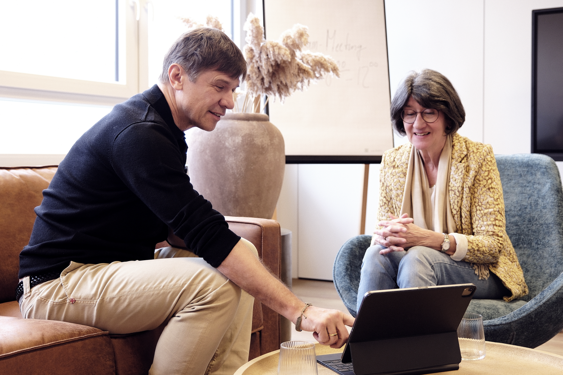 Zwei Personen sitzen in einem gemütlichen Raum, während sie an einem Tablet arbeiten; der Mann zeigt auf den Bildschirm, während die Frau aufmerksam zuschaut. Im Hintergrund ist ein Flipchart mit der Aufschrift "Meeting" und eine große Vase mit Trockenblumen zu sehen.