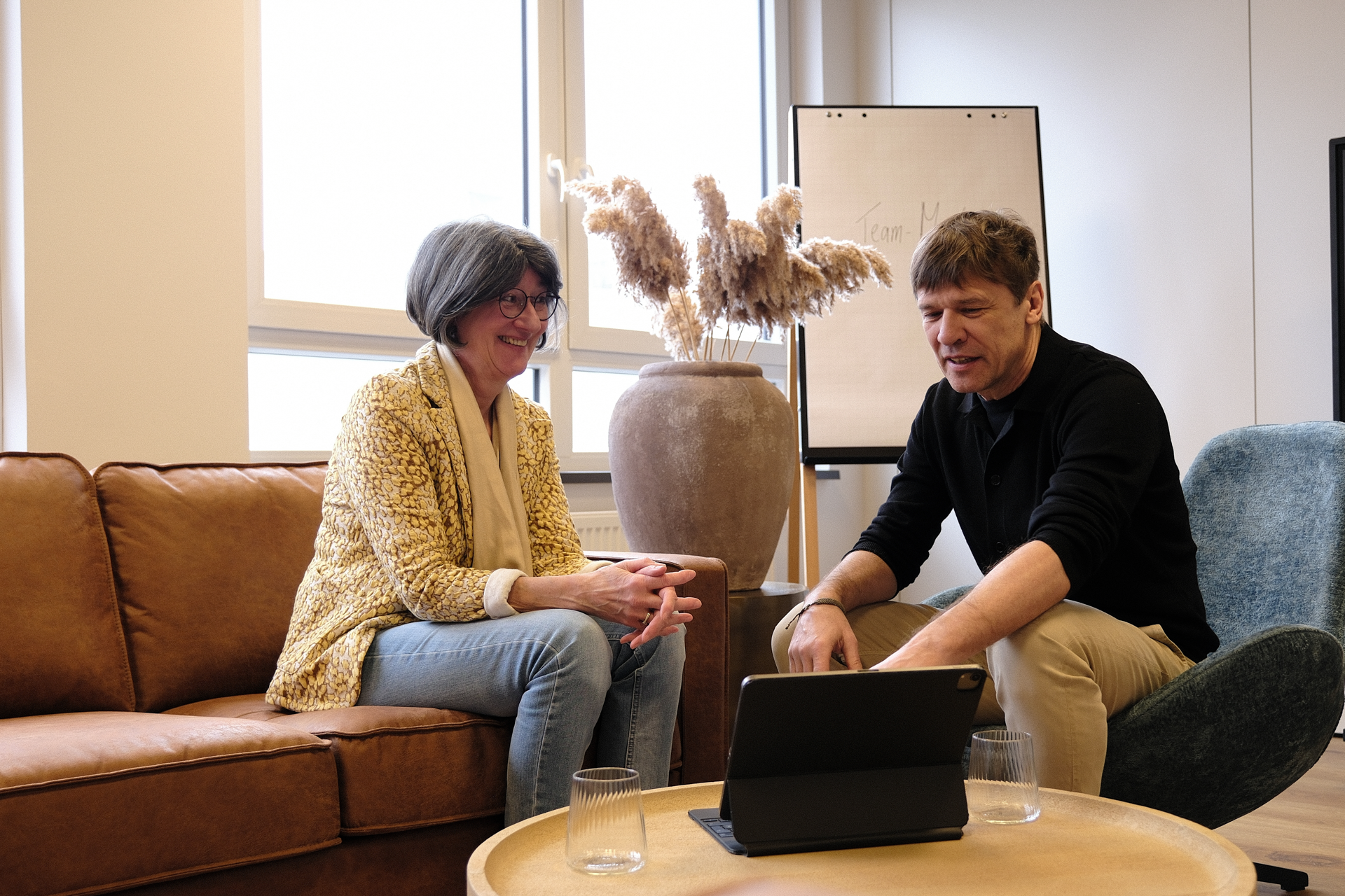 Eine Frau mit Brille und einer gelben Jacke sitzt auf einem Sofa und lächelt, während sie mit einem Mann spricht, der auf einem Tablet schaut. Im Hintergrund sind ein großes Fenster und ein Pflanzenelement in einer Vase zu sehen.