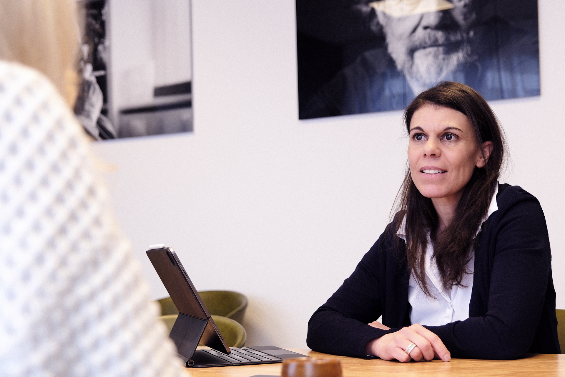 Eine Frau mit langen, dunklen Haaren sitzt an einem Tisch und diskutiert, während sie eine andere Person ansieht. Im Hintergrund sind schwarz-weiße Porträtfotos an der Wand zu sehen, und auf dem Tisch steht ein Tablet.