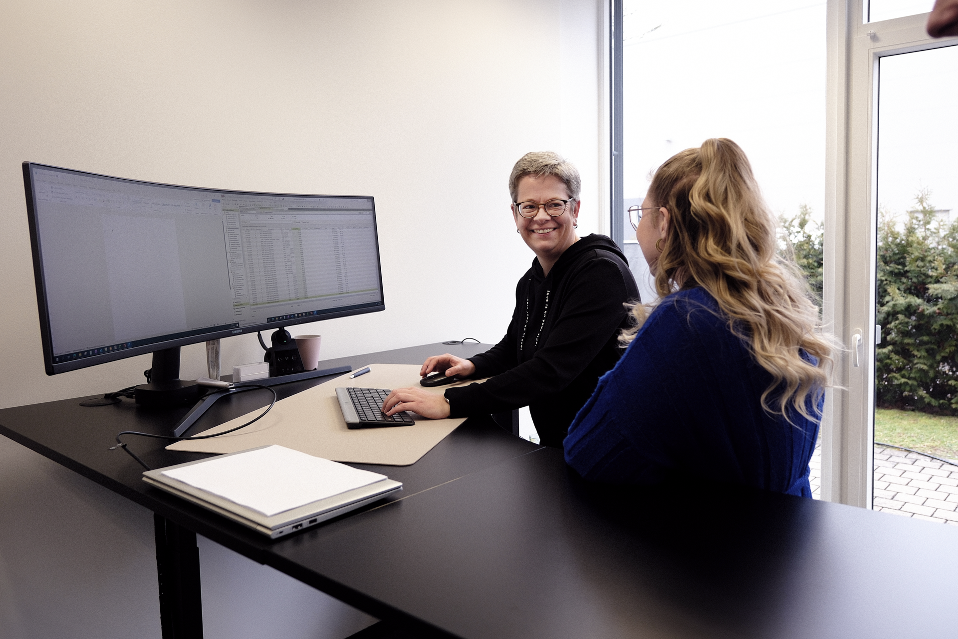 Zwei Frauen sitzen an einem Stehpult und arbeiten gemeinsam am Computer, während sie sich unterhalten. Auf dem Bildschirm sind Tabellenkalkulationen zu sehen. Im Hintergrund sind große Fenster mit Blick auf eine grüne Hecke.