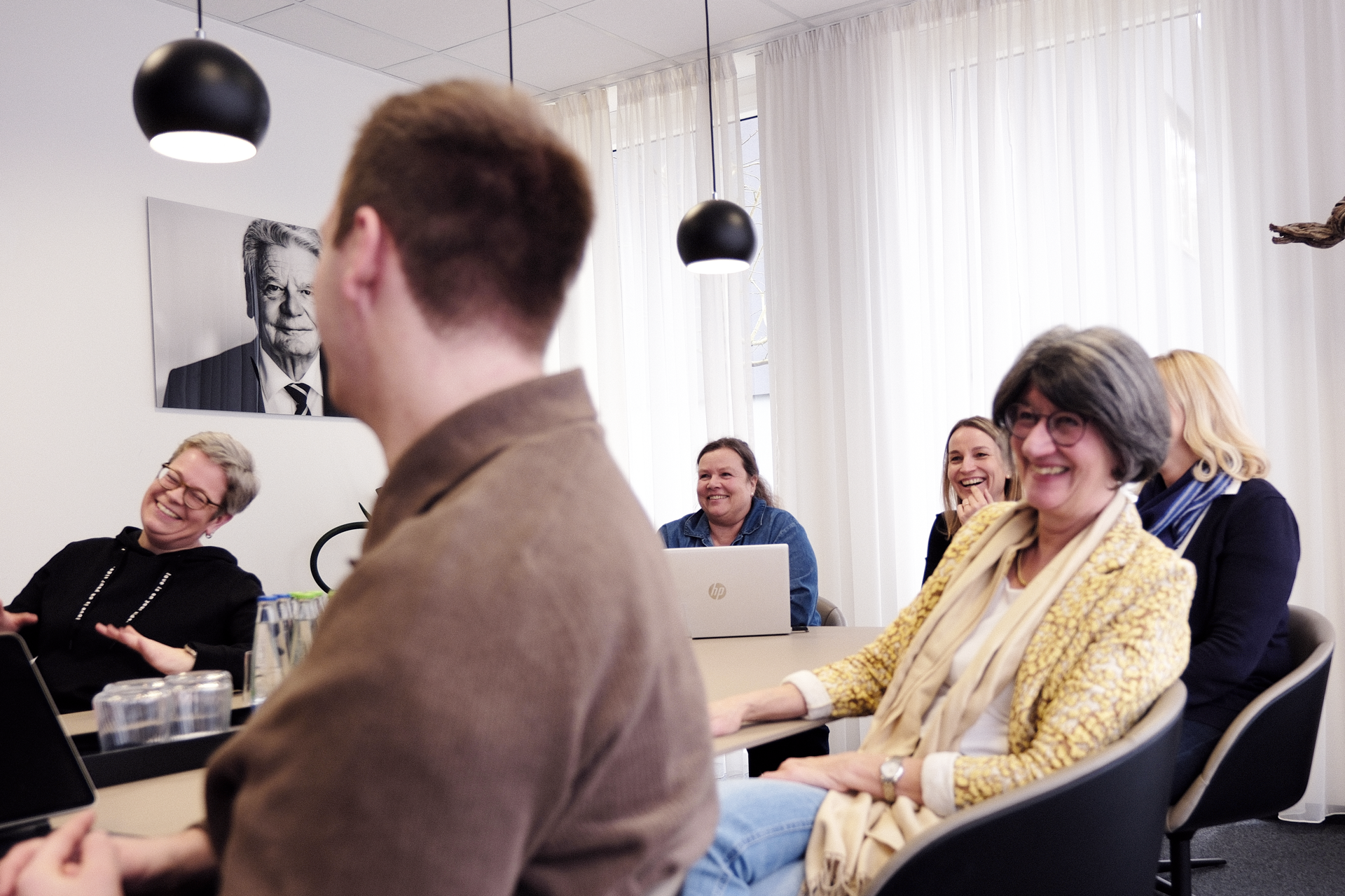 In einem modernen Büroraum sitzen fünf Personen an einem Tisch, die entspannt lachen und miteinander interagieren. An der Wand hängt ein Porträt eines älteren Mannes. Jeder Teilnehmer hat einen Laptop vor sich. Die Atmosphäre wirkt freundlich und informell.
