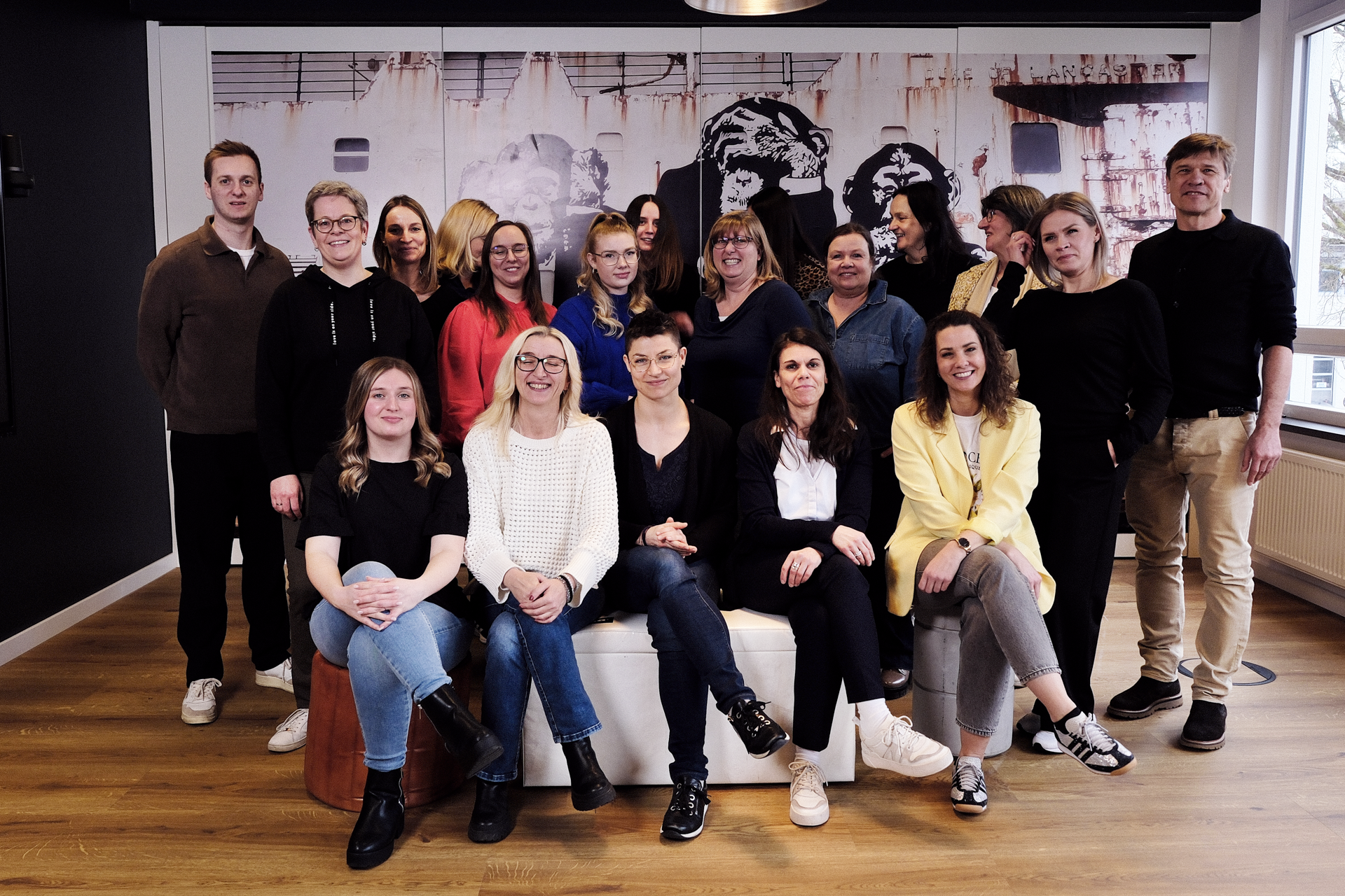Eine Gruppe von 21 Personen, die in einem modernen Büro steht und sitzt, posiert lächelnd vor einer muralen Wand mit einem künstlerischen Aufdruck. Die Personen sind unterschiedlich gekleidet und scheinen eine diverse Gruppe zu sein.