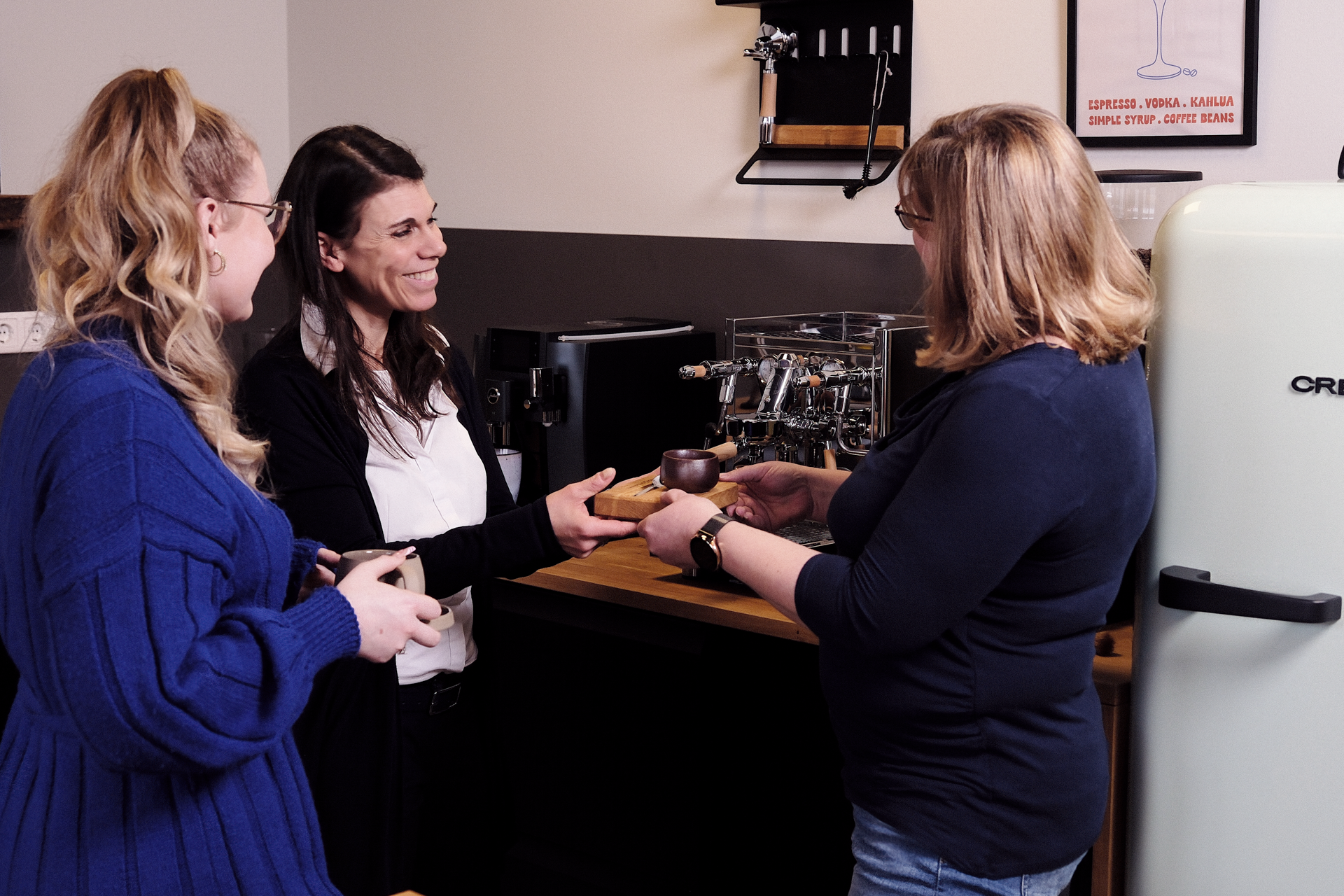 Drei Frauen stehen in einer modernen Küche. Eine Frau überreicht einer anderen einen Becher, während eine dritte Frau lächelnd zuschaut. Im Hintergrund sind eine Kaffeemaschine und ein Kühlschrank zu sehen.