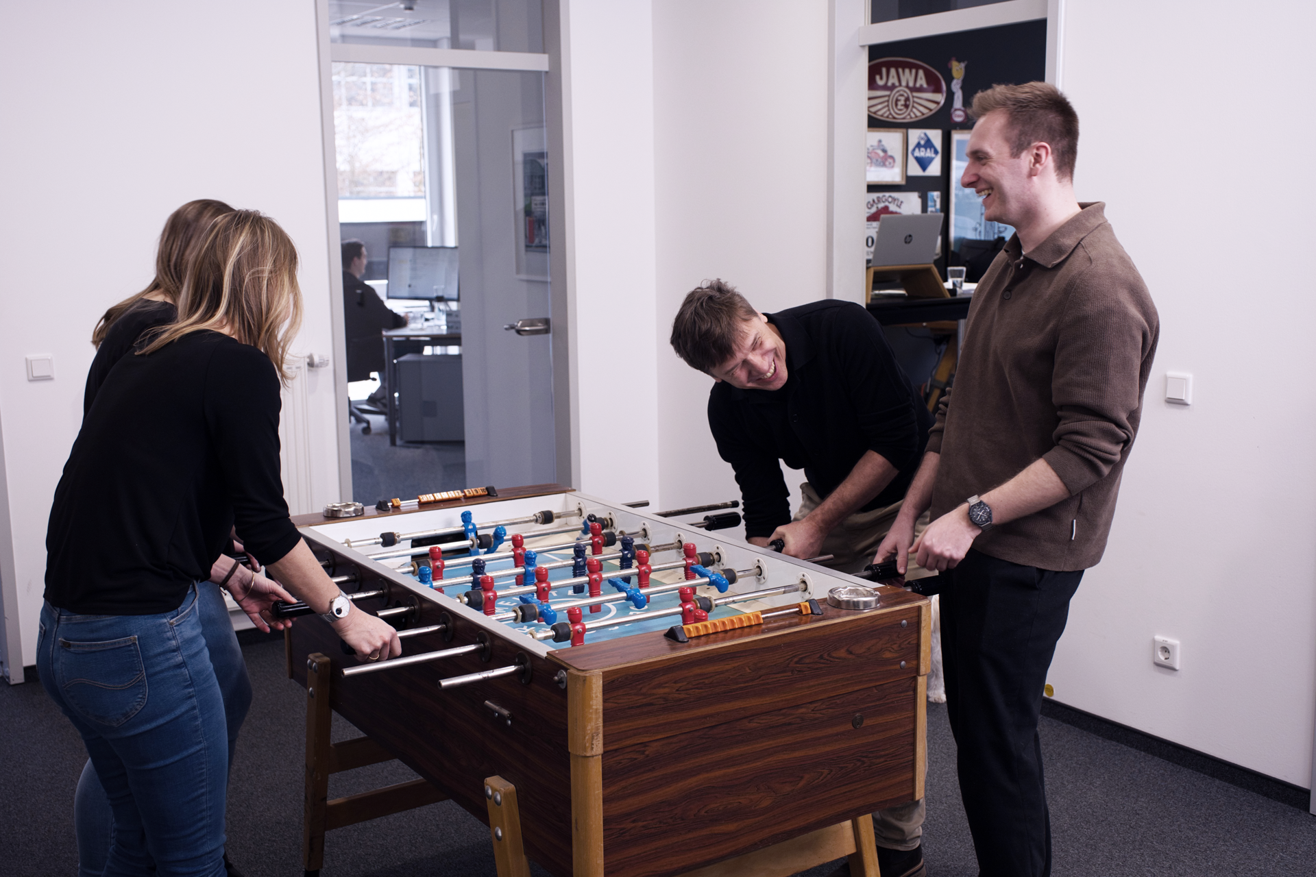 Vier Personen spielen gemeinsam an einem Tischfußballtisch in einem modernen Büro. Zwei Frauen und zwei Männer stehen um den Tisch herum und lächeln, während sie aktiv am Spiel teilnehmen. Im Hintergrund sind Arbeitsplätze und ein Computer sichtbar.