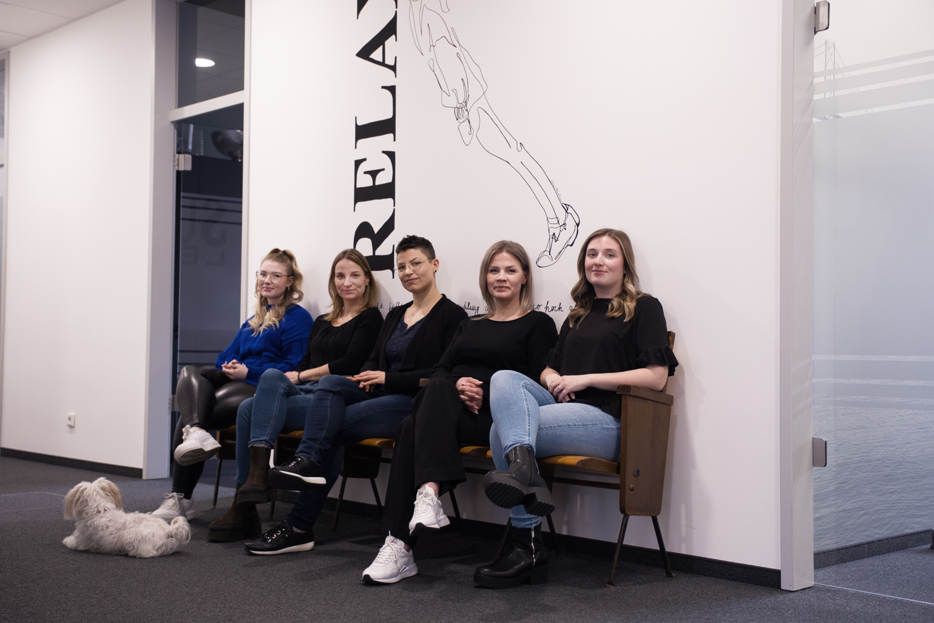 Fünf Frauen sitzen an einer Wand mit Wandgrafik, während ein kleiner Hund auf dem Boden liegt; alle tragen schwarze Oberteile und lächeln in die Kamera.
