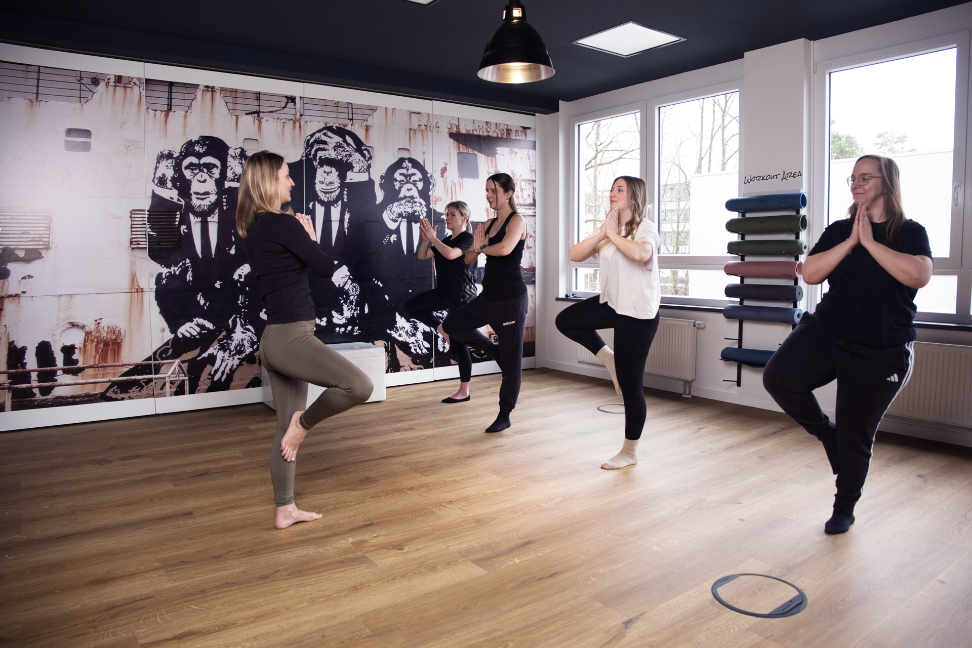 Fünf Frauen praktizieren in einem Raum mit einem Wandbild von Affen Yoga, wobei sie in einer Baum-Pose stehen, während sie die Hände vor der Brust zusammenführen.