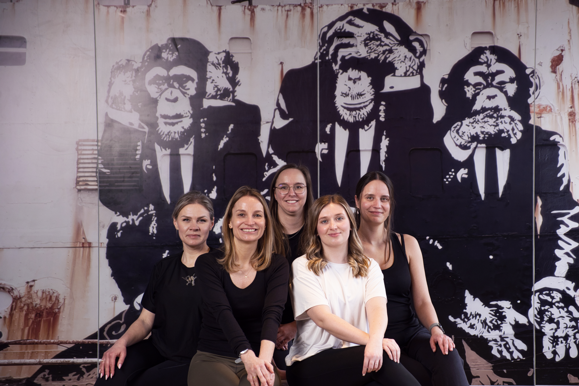Sechs Frauen posieren in lässiger Kleidung vor einer Wand mit einem großen, grafischen mural von Affen in Anzügen.