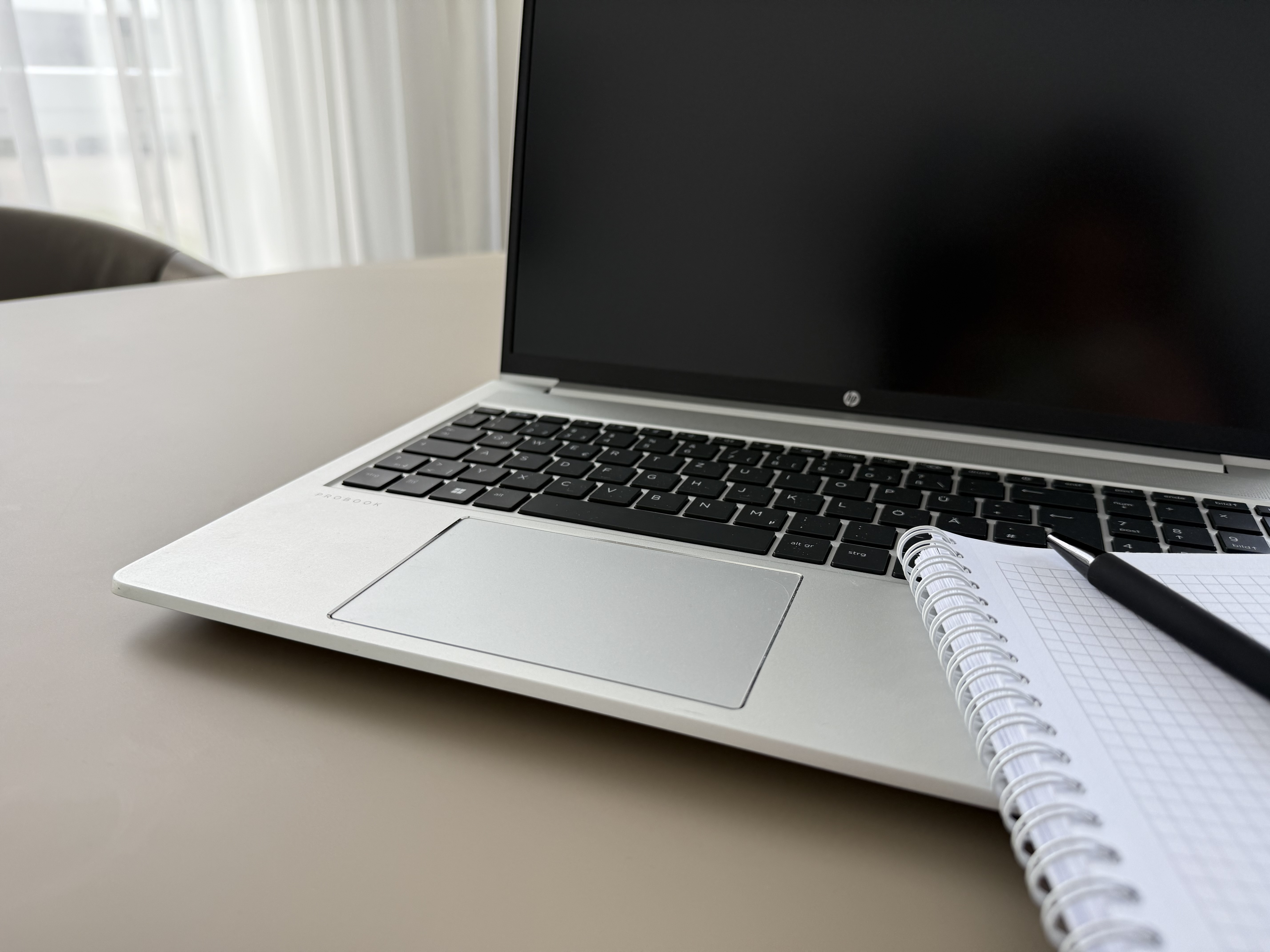 Ein Laptop steht auf einem Tisch neben einem Notizblock und einem Stift. Der Bildschirm des Laptops ist schwarz und der Hintergrund zeigt helle Vorhänge.