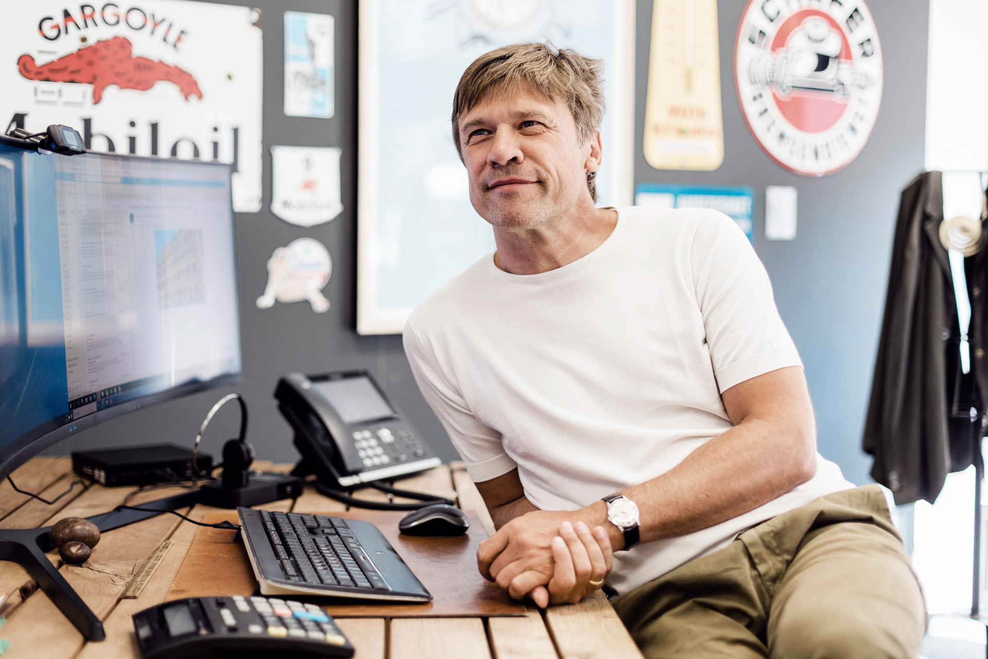 Ein Mann mit kurzem, braunem Haar sitzt entspannt an einem Holztisch, lächelt und schaut zur Seite. Vor ihm stehen ein Computerbildschirm und eine Telefontastatur, während im Hintergrund verschiedene bunte Wanddekorationen zu sehen sind.