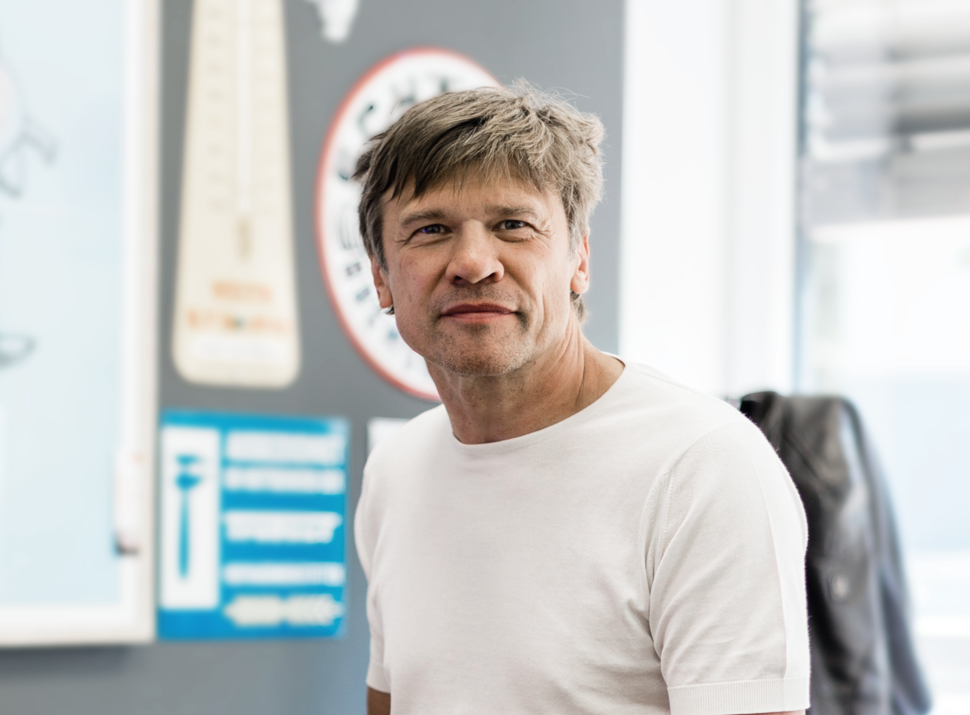 Ein Mann mit kurzen, grauen Haaren trägt ein weißes T-Shirt und lächelt, während er in einem hellen Raum steht. Im Hintergrund sind verschiedene bunte Schilder und ein Thermometer zu sehen.