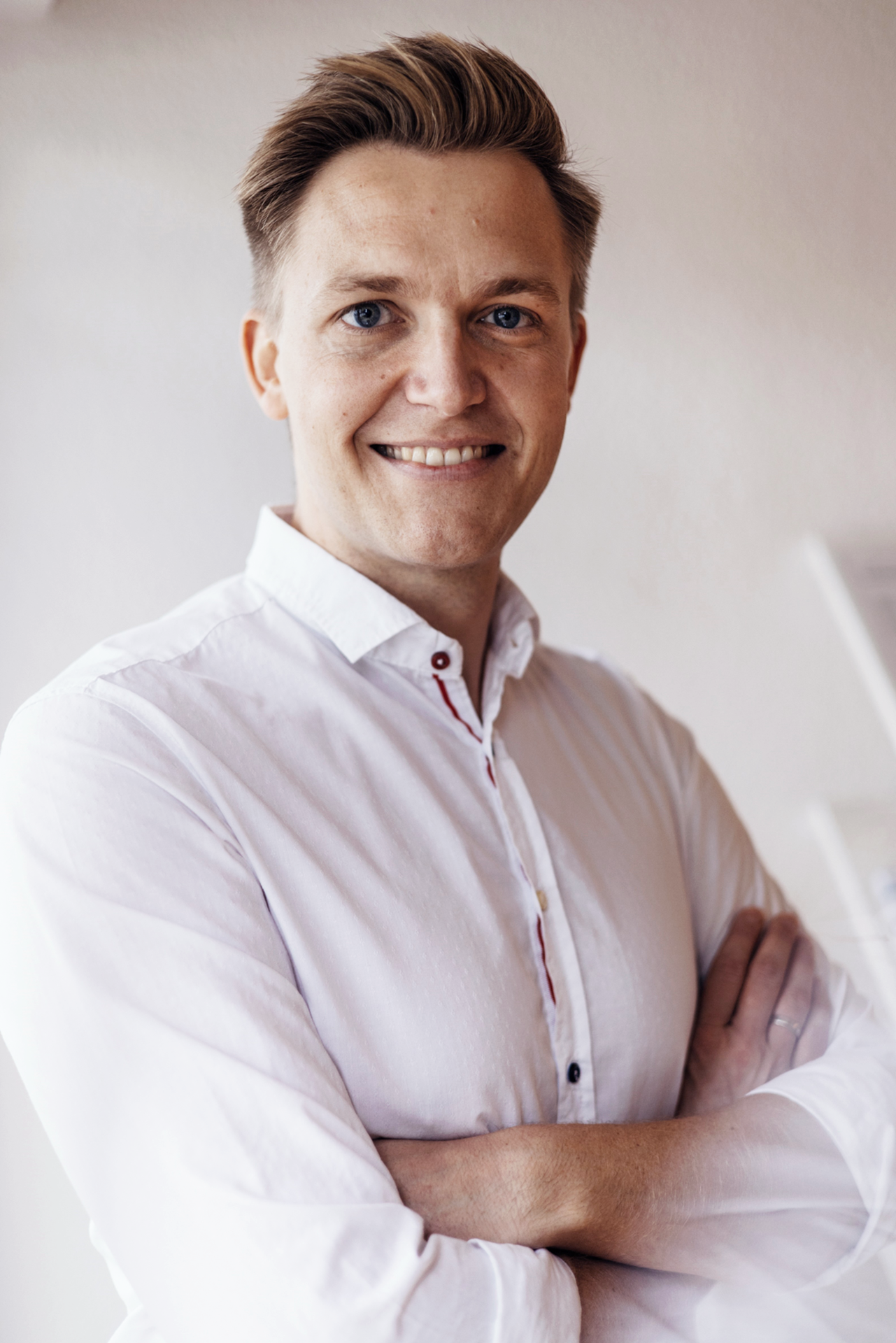 Ein lächelnder Mann mit blonden Haaren und einem weißen Hemd steht mit verschränkten Armen vor einer hellen Wand.