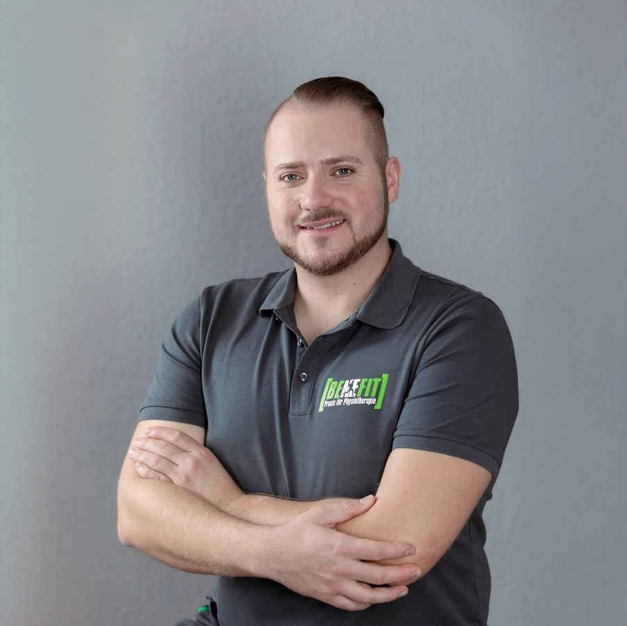 Ein lächelnder Mann mit kurzen, braunen Haaren und einem gepflegten Bart steht mit verschränkten Armen vor einer grauen Wand. Er trägt ein dunkelgraues Polo-Shirt mit dem Logo "Be-Fit" darauf.
